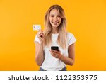 Portrait of a happy young blonde girl showing plastic credit card while holding mobile phone isolated over yellow background