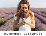 Photo of caucasian young woman in dress holding bouquet of flowers while walking outdoor through lavender field in summer