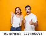 Excited beautiful couple wearing white t-shirts standing isolated over yellow background, showing thumbs up