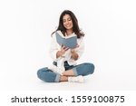Beautiful happy young brunette woman wearing casual clothing sitting on a floor with legs crossed isolated over white background, reading a book