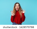 Image of joyful caucasian woman holding cellphone and gesturing ok sign while winking isolated over blue background