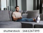 Image of a handsome cheerful young man indoors at home on sofa using laptop computer.