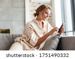 Image of happy nice woman in pajamas smiling and using cellphone while sitting on sofa at living room