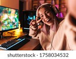 Image of excited girl taking selfie photo and gesturing peace sign while playing video game on computer indoors