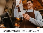 Young white waiter man wearing apron standing in front of computer and holding client receipt in cafe