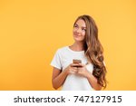 Photo of cheerful cute beautiful young woman chatting by mobile phone isolated over yellow wall background. Looking aside.