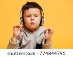 Photo of little concentrated boy child sitting on floor isolated over yellow background with eyes closed listening music with headphones.
