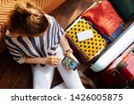 Small photo of Upper view of stylish woman in white pants and striped blouse in the modern house in sunny summer day book flight on phone near open travel suitcase.