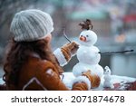 Seen from behind middle aged woman with mittens in a knitted hat and sheepskin coat making a snowman outside in the city park in winter.