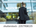 Seen from behind middle aged business woman near office building in black jacket with smartphone and briefcase walking.