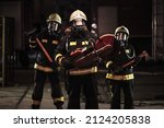 Small photo of Group of professional firefighters wearing full equipment, oxygen masks, and emergency rescue tools, circular hydraulic and gas saw, axe, and sledge hammer. Firetrucks in the background.