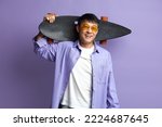 Smiling Man Holding Skateboard. Asian Guy Holding Longboard on Shoulder and Smiling while Posing Isolated on Violet Background 