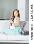 Small photo of Smiling small girl wearing blue dress sits on the couch witn a handsel and a glass in her hands