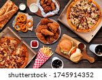 Small photo of Buffet table scene of take out or delivery foods. Pizza, hamburgers, fried chicken and sides. Above view on a dark wood background.
