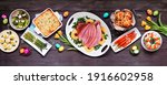 Small photo of Classic Easter ham dinner. Top view table scene on a dark wood banner background. Ham, scalloped potatoes, eggs, hot cross buns, carrot cake and vegetables.