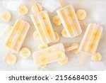 Healthy banana yogurt popsicles. Above view with scattered fruit slices and ice cubes over a bright white marble background.