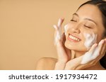 Beauty portrait of lovely young woman smiling with eyes closed while applying gentle foam facial cleanser isolated over beige background