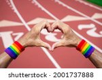 Small photo of Gay athlete wearing rainbow pride wristbands making love heart hands gesture against a red athletic track background