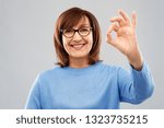 Small photo of gesture, vision and old people concept - portrait of smiling senior woman in glasses showing ok hand sign over grey background