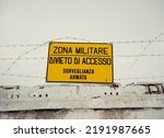 A yellow sign with the black text, in Italian: military zone, access forbidden. On top of a wall with barbed wire.

