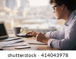Businessman using calculator on wooden office desktop with laptop, coffee cup, smartphone and business reports. Blurry city background