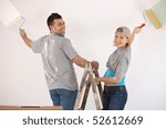 Portrait of smiling couple standing on ladder, painting wall together with paint roller.