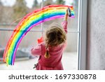 Adoralbe little toddler girl with rainbow painted with colorful window color during pandemic coronavirus quarantine. Child painting rainbows around the world with the words Let's all be well.