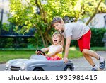 Two happy children playing with big old toy car in summer garden, outdoors. Kid boy pushing and driving car with little toddler girl, cute sister inside. Laughing and smiling kids. Lovely family.