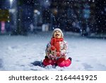 Adorable little toddler girl walking outdoors in winter. Cute toddler during strong snowfall on evening. Child having fun with snow. Wearing warm baby colorful clothes and hat with bobbles.