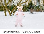 Happy little baby girl making first steps outdoors in winter through snow. Cute toddler learning walking. Child having fun on cold snowy day. Wearing warm baby pink clothes snowsuit and bobbles hat.