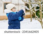 Little girl with glasses feeds birds on winter. Happy smiling preschool child hanging selfmade bird seed heart on tree. On cold snowy winter day.