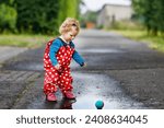 Small photo of Little toddler girl wearing rain boots and trousers and walking during sleet, rain on cold day. Baby child having fun with jumping in a puddle. Active outdoors leisure and activity with little kids.