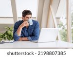 Small photo of Candid shot of careworn businessman sitting at desk with a laptop with hand on temple and working. Professional man drinking morning coffee. Home office.