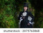 Small photo of Historical reconstruction of the late 19th and early 20th centuries. Elegant brunette lady in a strict black dress posing in an autumn park. Historical makeup and hairstyle.
