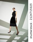 Small photo of Portrtait of a beautiful female fashion model in elegant tight black dress and stylish sunglasses posing in the studio on a white background with shadows. Full length shot.