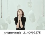 Small photo of Theater of life. The unfortunate girl stands with her eyes raised to the sky and hands clasped in prayer, surrounded by masks. Hypocrisy. Studio photo on a white background.