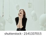Small photo of A girl in black clothes screams furiously, surrounded by white masks. Human roles. Hypocrisy. Mental disorders. Studio photo on a white background.