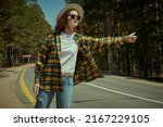 A beautiful girl traveler stands by a pine forest trying to catch a car on the road. Summer vacation outdoors. Tourism, hitchhiking.
