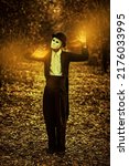 Small photo of Mysterious male mime in a white carnival mask, bowler hat and black tailcoat stands in an old autumn park with a mystical light in his hands. Halloween. Mystical horror novel. Vintage style.