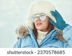 Winter fashion. Portrait of a fashionable girl posing in blue ski suit, white fur hat and stylish sunglasses  against a snowy winter landscape. Sport in the winter mountains. Copy space.