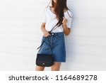 Details of trendy casual summer or spring outfit. Woman wearing blue denim mini skirt, white tshirt, small black cross body bag standing near white roller door. Everyday look. Street fashion. No face.