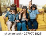 Three young women, two Caucasian and one Brazilian, aged 20-30, sitting on a couch in a gaming lounge, wearing VR headsets and using hand tracking technology. Casually dressed.