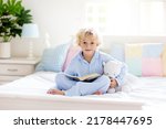 Child reading book in bed in white sunny bedroom with window. Children read books. Kids room. Little blond curly boy in pajamas at home. Bedding and sleepwear for nursery. Kid with toy.