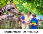 Happy family, young mother with two children, cute laughing toddler girl and a teen age boy feeding giraffe during a trip to a city zoo on a hot summer day
