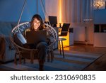 Woman enjoying leisure time at home, sitting in hanging chair in living room, using laptop computer, chatting online with friends and relaxing late at night