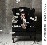 Small photo of Happy and shoutting arrogant rich kid boy millionaire sits with a bundle of money dollars cash in big luxury armchair while bills falling down from the sky. Money rain