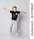 Small photo of Playful frolic blond kid boy in sunglasses, black t-shirt with dinosaur print and gray pants jumps with hands spread wide, has fun over gray background