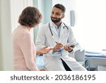 Small photo of Indian male doctor consulting senior old patient filling form at consultation. Professional physician wearing white coat talking to mature woman signing medical paper at appointment visit in clinic.