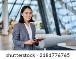Small photo of Smiling young Asian business woman leader holding digital tablet standing in office. Professional executive manager or saleswoman using corporate technology looking at camera. Portrait