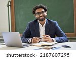 Happy young indian arabic businessman professional coach, teacher or university professor wearing suit looking at camera sitting at work desk in classroom office posing for portrait at workplace.